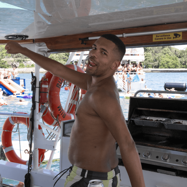 A man in swim trunks stands on a boat, pointing towards something off-camera. He is near a grill and a can of beer. Other boats float nearby, and people are enjoying the water behind him. The scene feels relaxed and summery, capturing the essence of a perfect beach day.