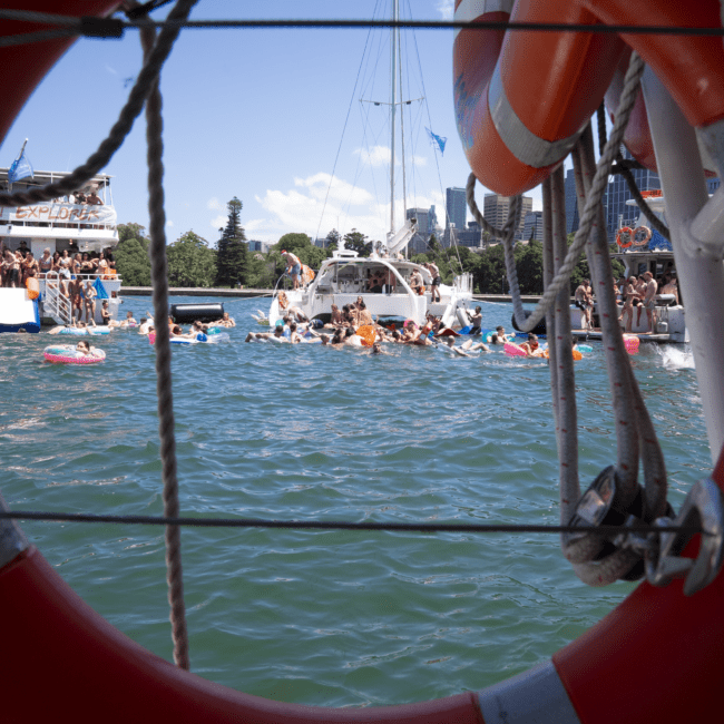 View through an orange life preserver showing a vibrant scene on the water with people swimming and socializing near boats. The background displays a clear sky and charming buildings along the waterfront. The image captures the essence of summer fun and camaraderie, evoking a sense of joy and relaxation.