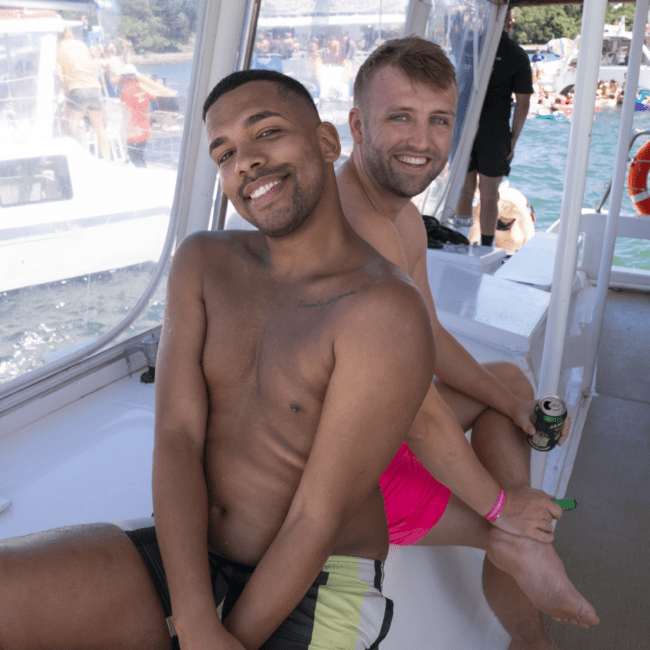 Two men sit on a boat holding drinks, smiling at the camera. One is shirtless with black and green swim trunks, and the other wears pink shorts. The boat is lively, and other boats are visible on the water in the sunny background.