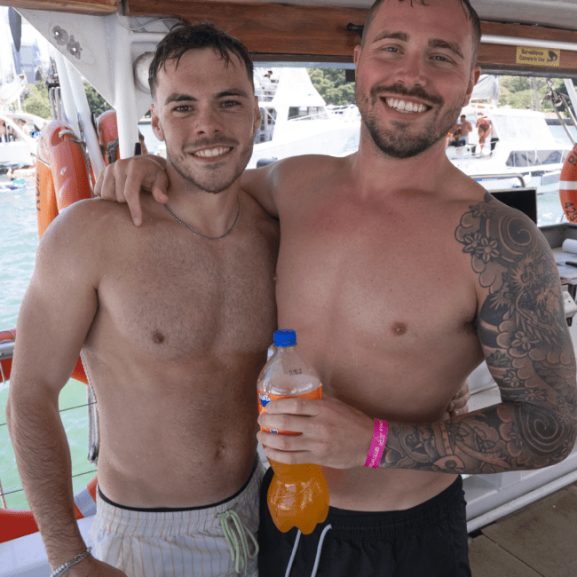 Two shirtless men pose on a boat with one man’s arm around the other’s shoulder. One has short hair and wears striped swim trunks, while the other, with a tattooed arm, holds a bottle of orange soda and wears black swim trunks. The background features sparkling waves and a clear blue sky.