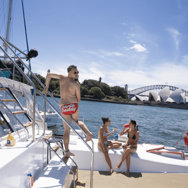 A man in red trunks is leaning on the railing of a boat near the Sydney Opera House and Harbour Bridge. Two women in swimsuits are sitting nearby, talking and holding a drink. The sky is clear and blue, with dolphins playfully swimming in the calm water below.