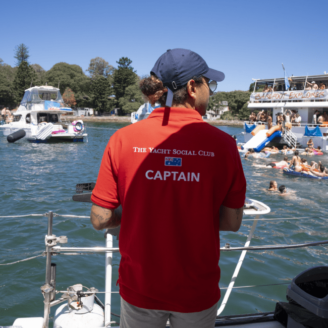 A person wearing a red polo shirt with "The Yacht Social Club" and "Captain" written on the back stands on a boat, overlooking a vibrant scene of people swimming and socializing on other yachts under the sunny sky.