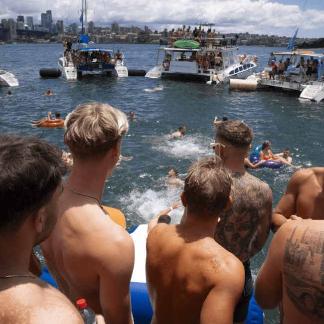 A group of shirtless men stand on a boat's deck, watching people swim near other anchored boats in a sunny harbor. The scene is lively, with several yachts and city buildings visible in the background under a blue sky with minimal clouds.