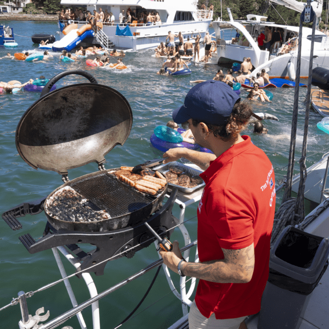 A person wearing a red shirt and a blue cap grills food on a barbecue placed on the deck of a boat. In the background, people are gathered on a floating platform and in the water, enjoying watersports. Other boats and a wooded shoreline are also visible.