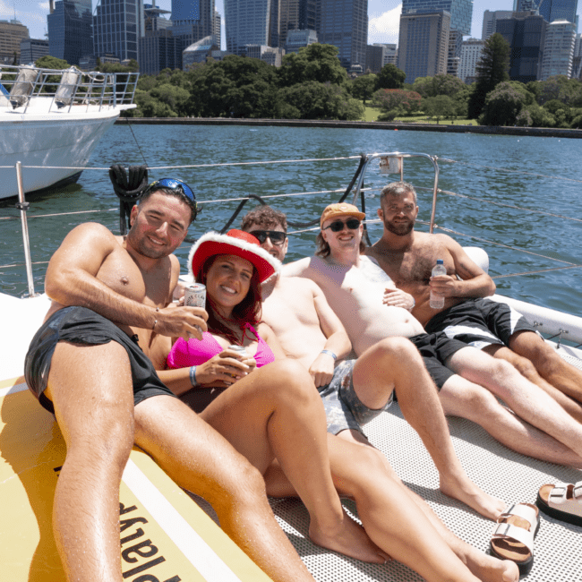 A group of people relax on the deck of a boat, basking in the sun with a city skyline in the background. Some are holding drinks and smiling while others lounge comfortably. The water and majestic city buildings create a picturesque backdrop to the serene scene on the boat.