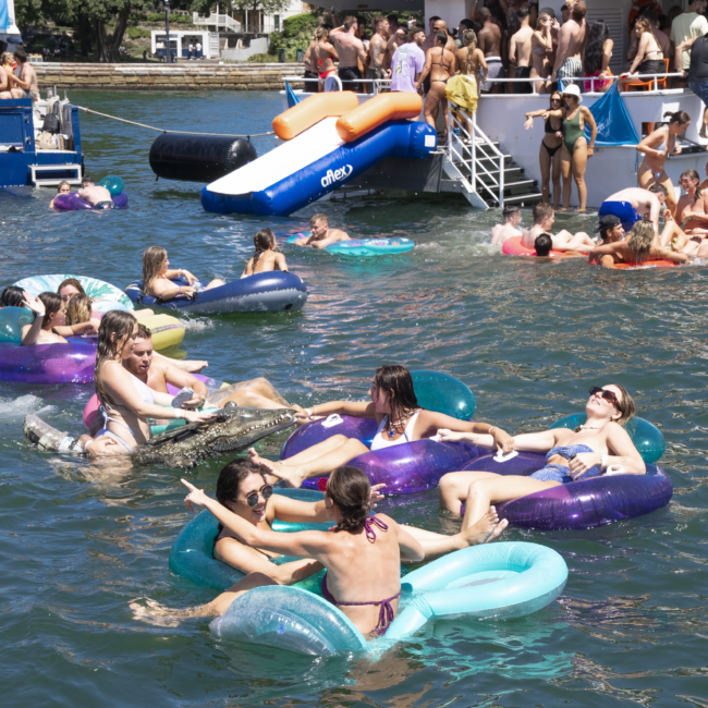 People in colorful swimsuits are floating on inflatable rings and rafts in the water near a boat named "Barefoot Explorer." The boat is crowded with more people, and an impressive inflatable slide extends from the boat into the water. Trees and a building are visible in the background, adding to the scenic view.