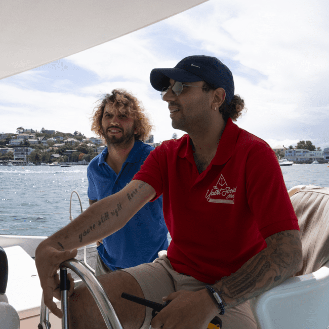 Two men are on a boat, sailing. One is steering the wheel, wearing a red shirt and sunglasses, with visible tattoos on his arms. The other man stands beside him, smiling with shoulder-length hair and a blue shirt. In the background, a coastal town meets the reflective water under the clear sky.