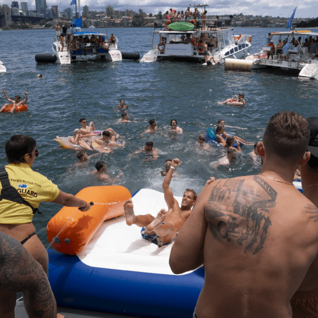 A vibrant group of people enjoying a sunny day on the water. Some are swimming, while others are on floating devices. A man slides down an inflatable slide into the water with joy. Several boats are anchored in the background, with lifeguards ensuring everyone's safety and adding to the lively atmosphere.