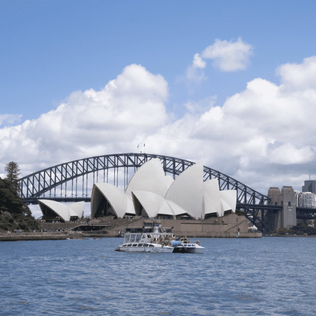 A boat cruises on the water in front of the iconic Sydney Opera House with its distinctive white sails, while the steel arch of the Sydney Harbour Bridge stands proudly against a blue sky with scattered clouds.