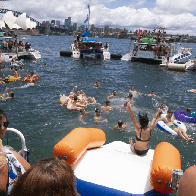 A lively scene on the water with numerous people swimming, floating on inflatables, and on boats. A person splashes into the water from an inflated slide. In the background, kayakers and buildings, including the Sydney Opera House, are visible under a partly cloudy sky.