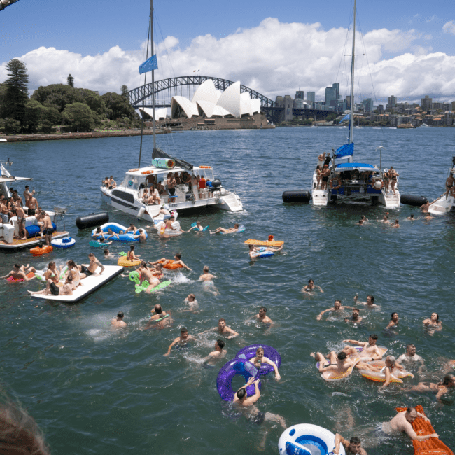 A lively scene of people swimming and floating on inflatable rafts in a body of water. Numerous boats are parked nearby, and in the background, the iconic Sydney Opera House and a bridge are visible against a blue sky with scattered clouds.