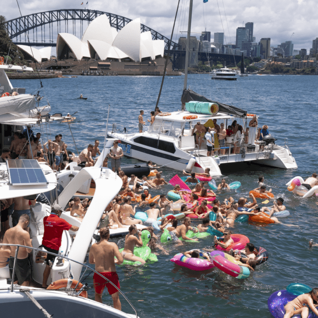 A lively scene of people celebrating on boats and colorful inflatable toys in the water. In the background, the Sydney Opera House and Sydney Harbour Bridge are visible against a clear, sunny sky. The vibrant waterfront adds to the excitement and festive atmosphere.