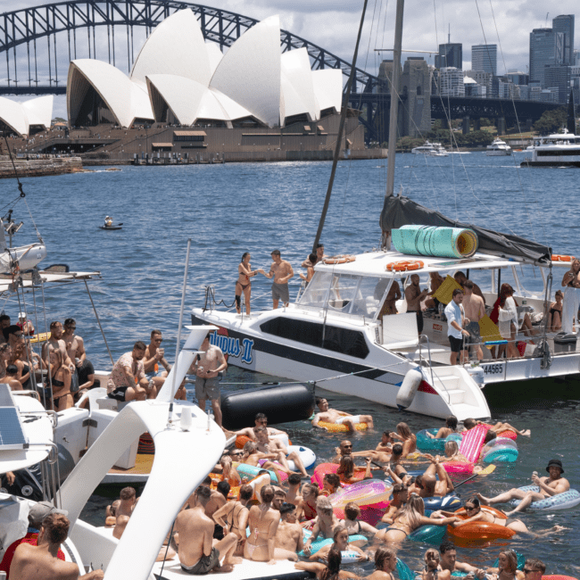 A large group of people enjoying a sunny day on the water next to boats, including a catamaran and a motorboat, with many floating on colorful inflatables. The iconic Sydney Opera House and Sydney Harbour Bridge are visible in the background against a clear blue sky, adding to the scenic charm.