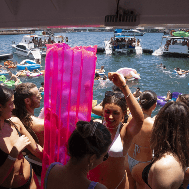 A large group of people gather on boats and in the water for a festive party. Several are on inflatable floats, many wearing colorful swimsuits. Drinks in hand, the atmosphere is lively and celebratory. The background features blue water shimmering from the sunshine and other boats dotting the scene.