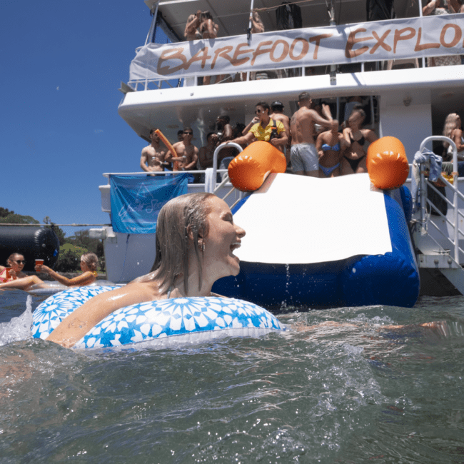 A woman smiles while floating on a blue and white inflatable tube in the water near a boat labeled "BAREFOOT EXPLORER." People on the boat and in the water enjoy the sunny day, some diving off the boat and interacting. The scene is one of pure bliss, a perfect day for barefoot adventure.