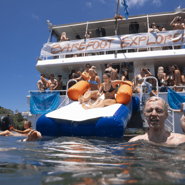 A group of people enjoys a sunny day on a double-deck boat named "Barefoot Explorer." Some are on the boat, while others dive into the crystal-clear water. A person is descending a slide into the water, and everyone seems to be having fun and socializing, taking in the picturesque scenery.