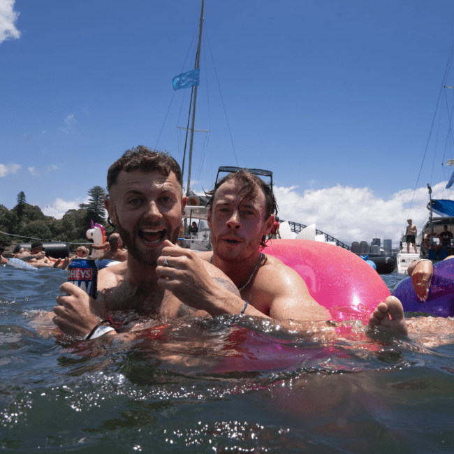 Two men are smiling and embracing in the water. One holds a can of drink. Behind them, other people are floating on inflatables, and sailboats are anchored nearby. The clear blue sky suggests a sunny day. Trees and cityscape are visible in the distance, creating a picturesque backdrop for this joyful scene.