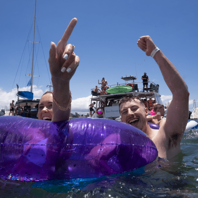 Two people in the ocean joyfully wave and cheer, surrounded by boats and clear blue sky. The person on the left points upward while inside a colorful inflatable tube, while the person on the right raises an arm in excitement. The background features more boats and happy people enjoying the day.