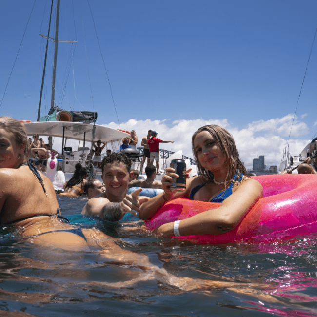 A group of people enjoying a sunny day at a beach party. Three individuals are in the foreground, floating on inflatable rafts in the water, with sailboats in the background. The sky is clear, and the atmosphere is lively and festive.