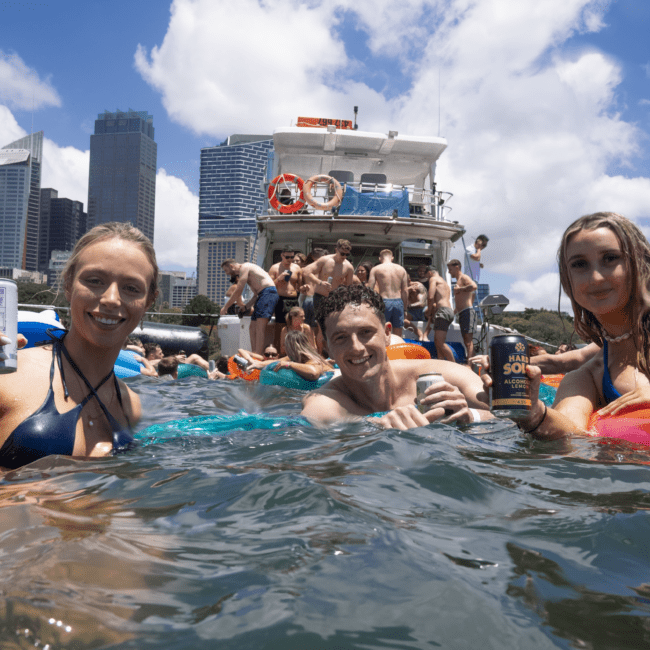 A group of friends enjoying a sunny day swimming in open water near a boat. Three people in the foreground smile at the camera, holding drinks, with a city skyline and clear blue sky in the background. They are surrounded by colorful inflatables and beach accessories.