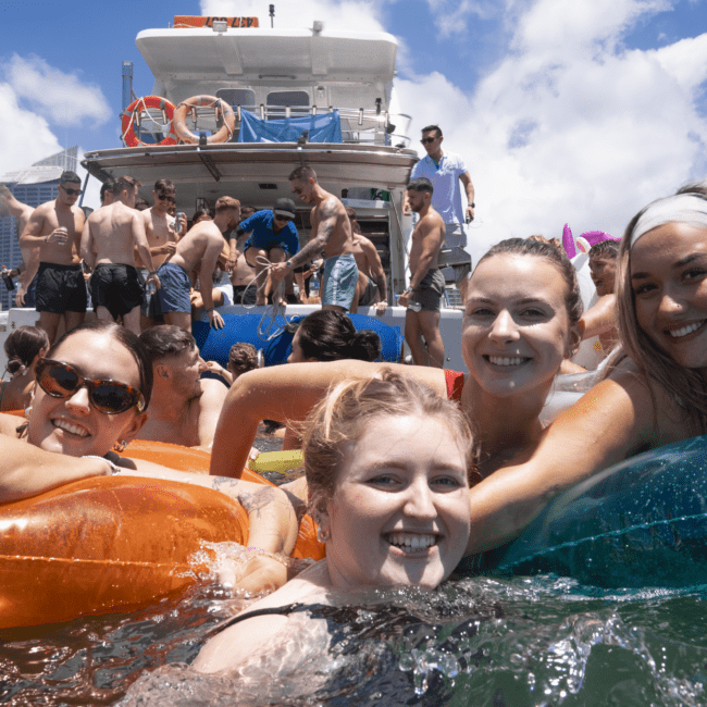 A lively group of friends enjoys a sunny day in the water, floating on colorful inflatables near a boat. A crowd is visible on the deck, with a cityscape and blue sky in the background. Everyone appears happy and relaxed, soaking up the vibrant atmosphere around them.