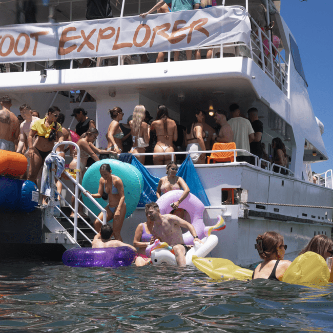 A group of people in swimsuits are enjoying a party on and around a large boat named "Boot Explorer." Many are on inflatable pool toys or lounging near the boat's steps, while others are chatting and socializing on the deck. The water is calm and clear, perfect for paddleboarding.