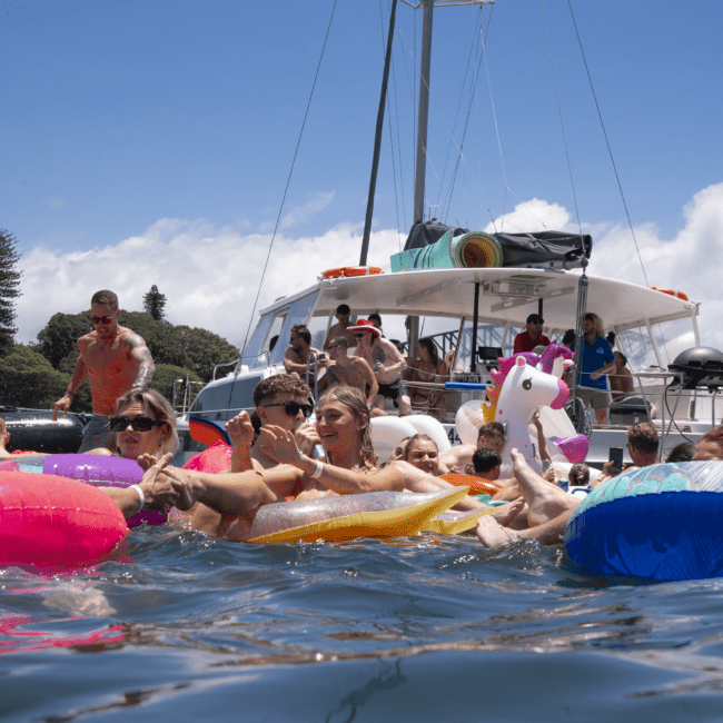 A group of people enjoying the water on colorful inflatable floats near a boat with a vibrant blue flag. The scene is lively with clear blue skies and trees in the background. Some individuals are laughing and interacting, creating a fun and festive atmosphere reminiscent of an aquatic festival.