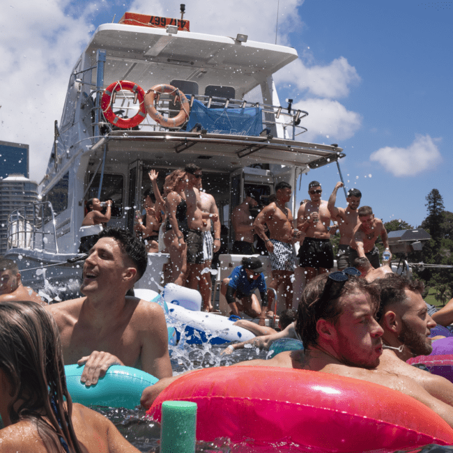 A lively boat party with people on board and in the water, surrounded by inflatable pool toys. The boat is anchored under a sunny sky with everyone enjoying themselves, splashing around, and socializing. The backdrop features a city skyline and some lush trees.