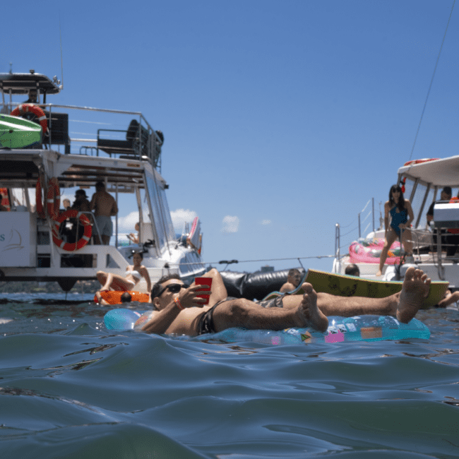 People are floating on inflatable loungers in the water, surrounded by boats. Others are on the sailboats relaxing and enjoying the sunny day. The sky is clear, and the atmosphere appears relaxed and leisurely.