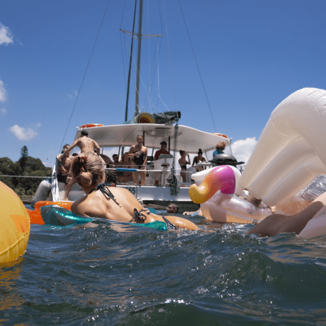 People are relaxing on various inflatable pool floats, such as a unicorn, in the water near a boat. The boat has several people on board, and a clear blue sky with some clouds is in the background. The colorful floats add vibrancy to the scene.