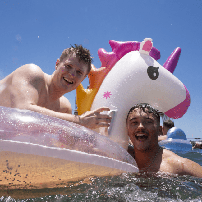 Two smiling individuals enjoy a sunny day in the water while holding large inflatable floats. One float resembles a glittery pool mat, and the other is shaped like a colorful unicorn. The clear blue sky enhances their joyful moment as they revel in the vibrant atmosphere.