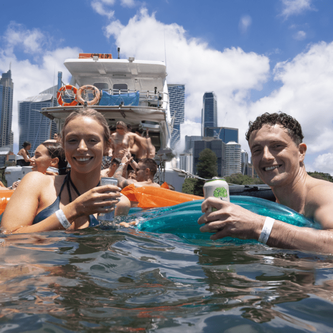 People enjoy a sunny day in a bustling city harbor, floating on inflatable tubes with refreshing cocktails in hand. Skyscrapers and blue skies are in the background, while a colorful boat with more people is docked nearby.