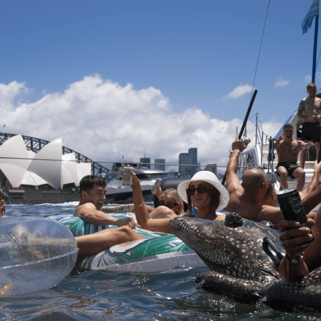 A group of people enjoy a sunny day on the water. Some are on inflatable floats, including a crocodile float. They are near a sailboat with the Sydney Opera House and Sydney Harbour Bridge visible in the background. The vibe is festive and relaxed, reminiscent of a joyful summer adventure.