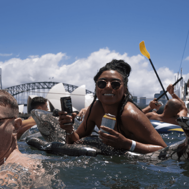 A woman in sunglasses and braids floats in water on a large inflatable snake, holding a can in each hand. Other people float nearby, and a bridge and white structure are visible in the background under a bright sky. The scene is reminiscent of various travel destinations designed for leisure.