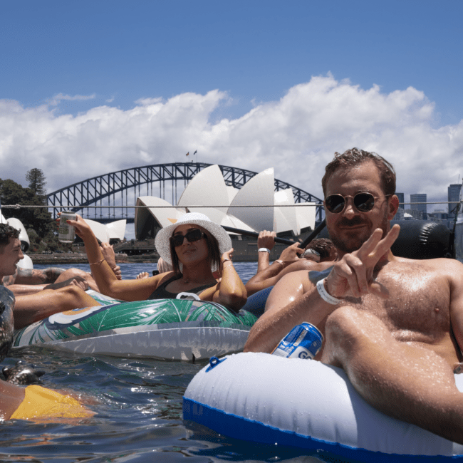 A group of people enjoy floating on inflatable tubes in water with the Sydney Opera House and Sydney Harbour Bridge in the background. One man donning sunglasses flashes a peace sign, while a woman wearing a hat smiles at the camera. It's a sunny day with scattered clouds, perfect for relaxation.