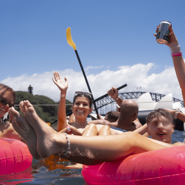 A group of people are joyfully floating on pink inflatable rafts in a body of water. One woman waves and smiles at the camera while holding a drink. Another person holds a paddle. A bridge and a famous landmark resembling the Sydney Opera House are visible in the background, adding to the scenic beauty.