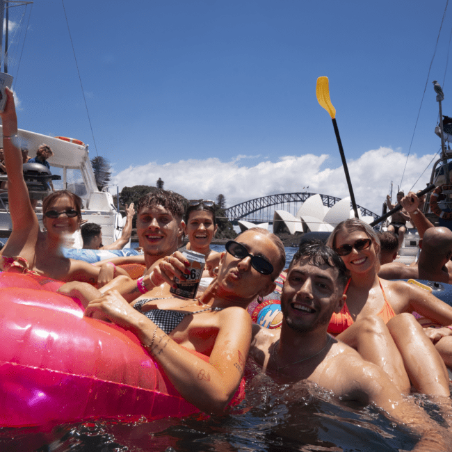 A joyful group of people in swimsuits is gathered on inflatable rafts in the water, smiling and posing for the camera. Boats are visible in the background, and kayaks add to the fun. The bright sun and clear sky indicate a beautiful day, with a prominent bridge seen in the distance.