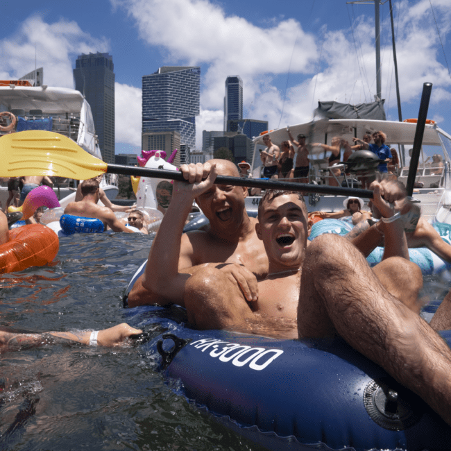 A group of people enjoying a sunny day on the water. Two men are at the center, smiling and holding up paddles while sitting in an inflatable raft. A yacht with more people can be seen in the background, with skyscrapers of a vibrant city skyline behind them. The scene captures a perfect urban adventure.