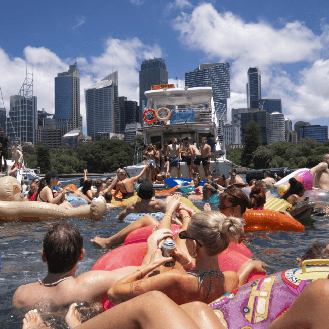 A large group of people enjoying a sunny day on a sparkling body of water, surrounded by numerous colorful inflatable floats and boats. They are relaxing, socializing, and drinking beverages. Skyscrapers and city buildings tower in the background under a blue sky with some scattered clouds.