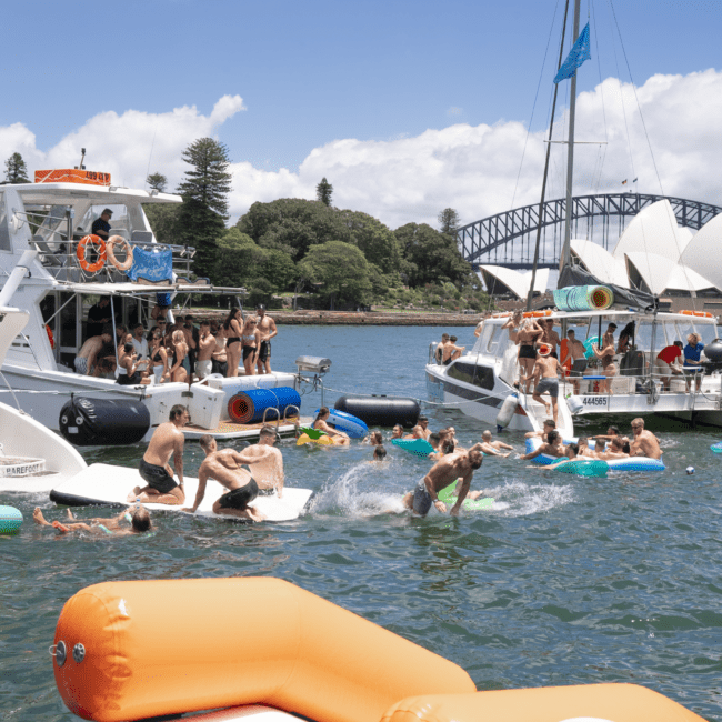 A lively scene of people enjoying a sunny day on the water, with several boats and a large inflatable raft in view. Some people are swimming, while others are kayaking. A prominent bridge and lush greenery are visible in the background.