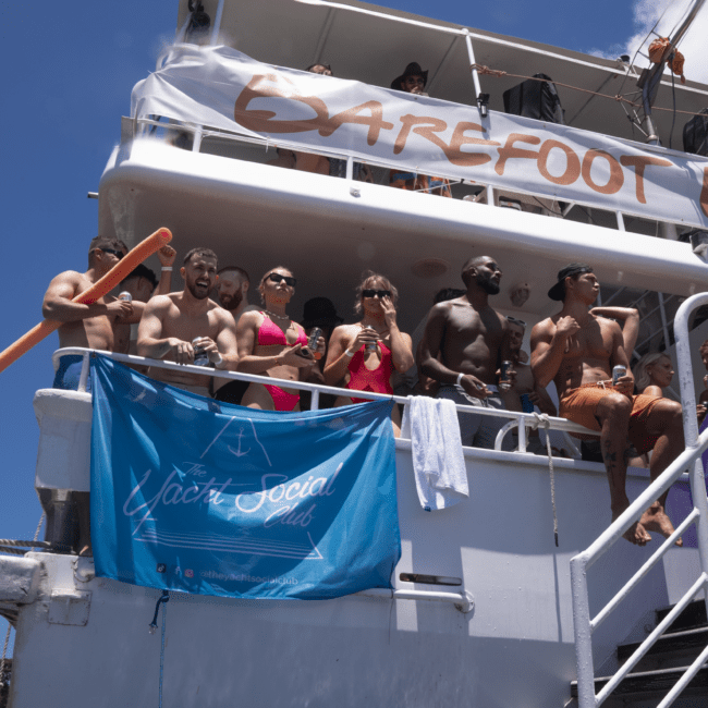 A group of people in swimwear are gathered on the deck of a boat labeled "BAREFOOT." They are enjoying a sunny day, with some holding drinks and chatting. A blue banner with "Yacht Social" is draped over the railing, and a pristine ocean stretches into the horizon under the clear blue sky.