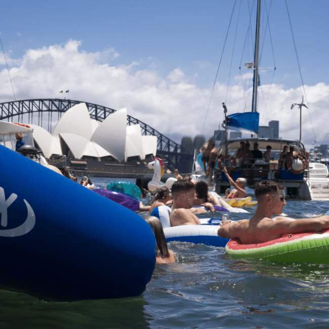 People are enjoying a sunny day on colorful inflatables in a body of water. In the background, there are boats, the Sydney Opera House, and the Sydney Harbour Bridge. The clear sky with a few clouds enhances the serene atmosphere.