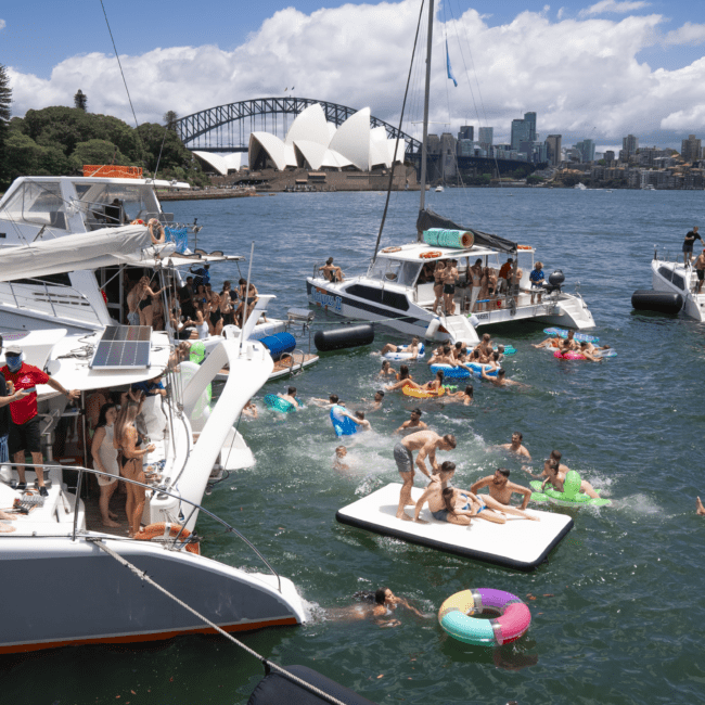 Boats are anchored in a sunny harbor where numerous people are swimming, floating on inflatables, and sunbathing on a large float. In the background, the iconic Sydney Opera House and Sydney Harbour Bridge complete the picturesque city skyline.