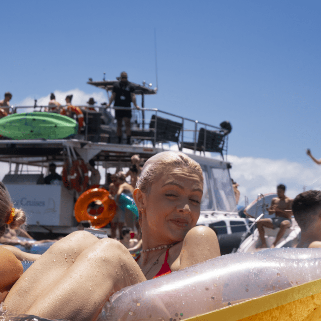 A person with light blonde hair is relaxing on a yellow float in the water, smiling with eyes closed. In the background, people are gathered on a boat under a sunny, clear blue sky. The boat features life rings and kayaks attached, adding to the scenic charm.