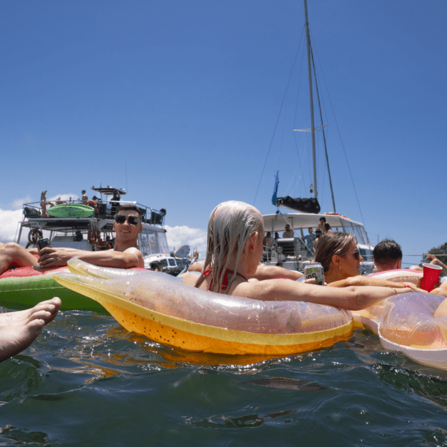 A group of people relax on inflatable floaties in the water, surrounded by boats. Feet in the foreground suggest someone is watching from their kayak. The sky is clear with a few clouds, and the scene radiates a joyful, leisurely vibe under the sun.