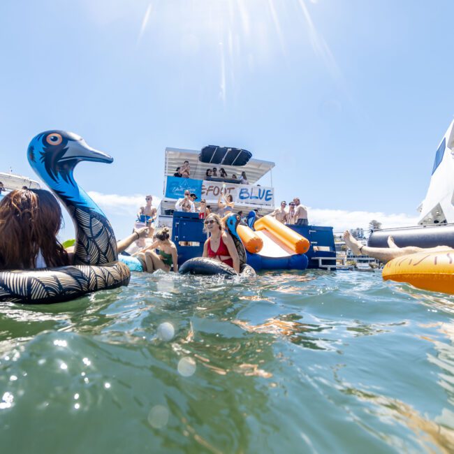 People enjoy a sunny day on a lake, floating on colorful inflatable toys and swimming near boats. The water is crystal clear under a bright blue sky. The scene is lively with social activities around a floating platform and kayaks anchored nearby.