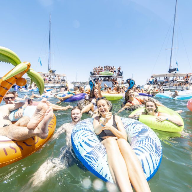 A group of people enjoying a sunny day on inflatable floaties in the water, surrounded by sailboats. The crowd appears cheerful and relaxed, with some holding drinks. Palm tree and donut-shaped inflatables are visible. The sky is clear and blue.