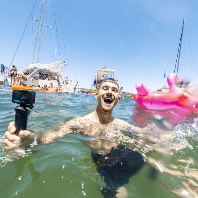 A man smiling while standing in water, holding a camera mounted on a grip stick. In the background, yachts and boats are filled with people relaxing, some on inflatable floats like a pink flamingo, under a vibrant blue sky. The lively water adds to the joyful atmosphere.