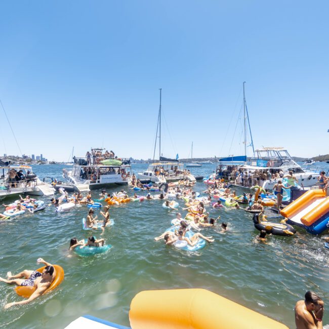 A lively scene on calm water with numerous people enjoying a sunny day. Some are swimming, while others relax on colorful inflatables. Several boats are anchored nearby, serving as gathering spots for their adventures. The sky is clear and bright, enhancing the festive atmosphere.