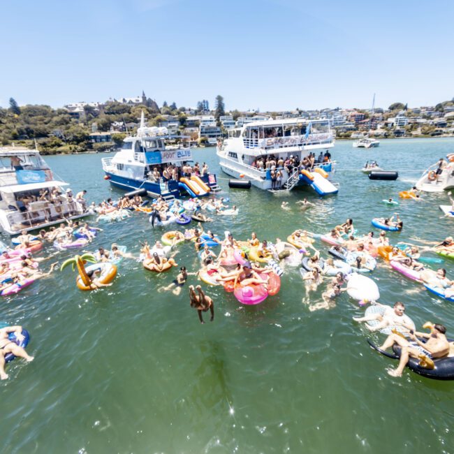 A large group of people are enjoying a sunny day on the water, lounging on various colorful inflatables. Several boats are anchored nearby, and live music equipment is set up on one boat. The backdrop includes a shoreline with greenery and houses on a hill.